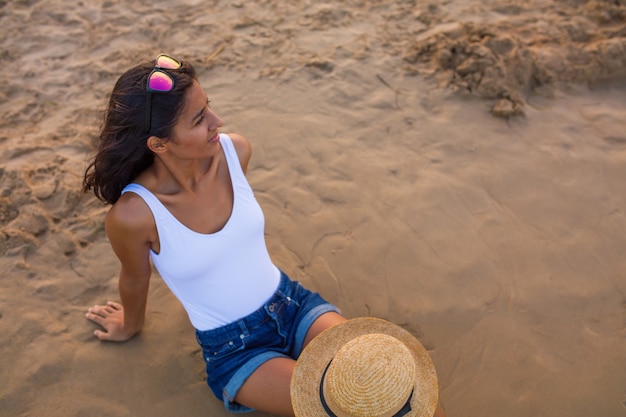 Foto giovane donna seduta sulla spiaggia con cappello e occhiali da sole