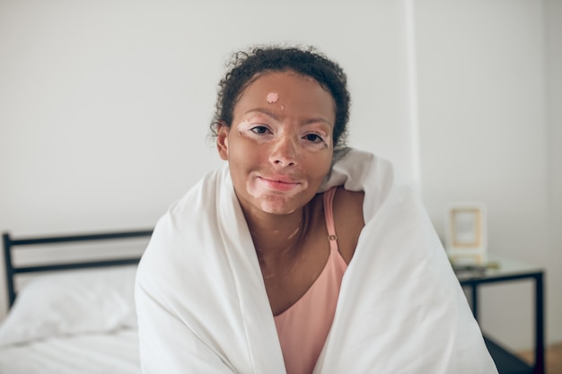 Young woman sitting in bed covered with blanket and feeling relaxed