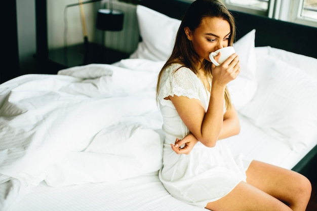 Молодая женщина, сидя на кровати и пить кофе после пробуждения