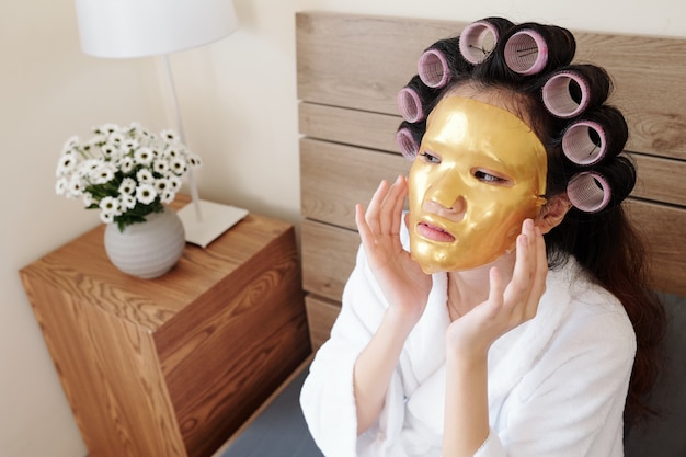 Young woman sitting on bed with hair rollers on her head and applying silicone sheet mask on face