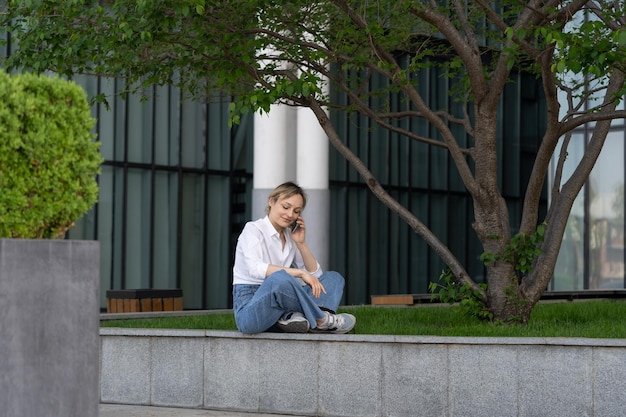 야외에서 휴대 전화로 이야기를 하고 있는 나무 아래 봄 공원의 벤치에 앉아 있는 젊은 여성