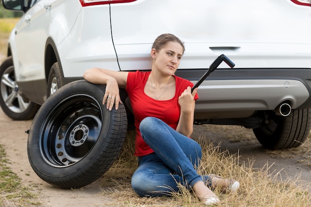 壊れた車の横に座って、パンクしたタイヤを交換しようとしている若い女性