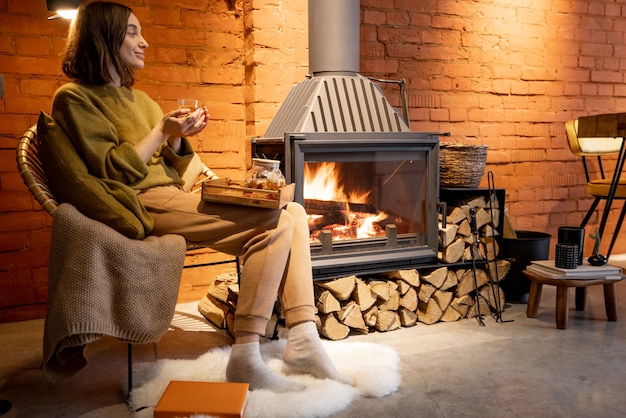 Young woman sitting by a burning fireplace, relaxing with a hot\
drink in cozy loft style interior during a winter time