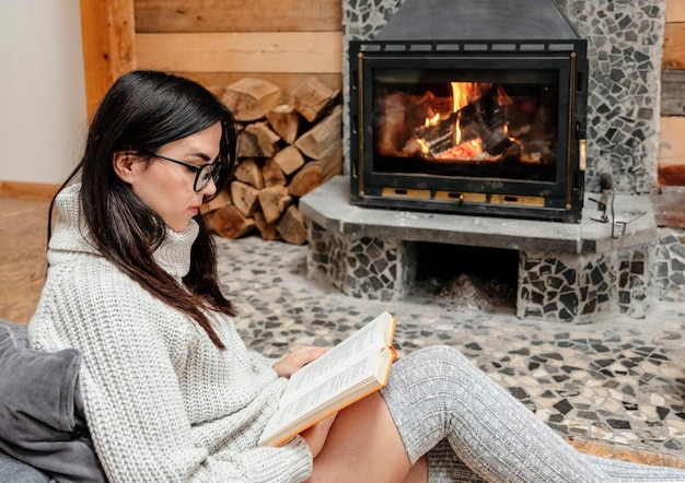 young woman sitting by fireplace in cozy cabin reading a book
