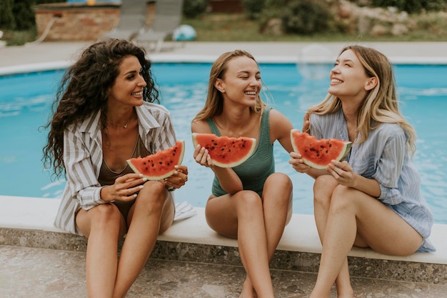 Young woman sitting on by the swimming pool and eating watermelon in the house backyard