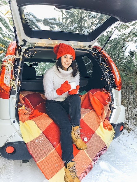 冬の雪の日に暖かいお茶を飲んで車のトランクに座っている若い女性