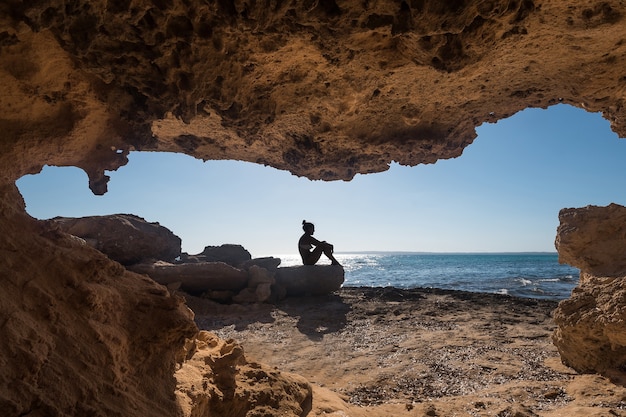 Young woman sitting next to a cave of a rock formation in front of the sea in Formentera in Spain.