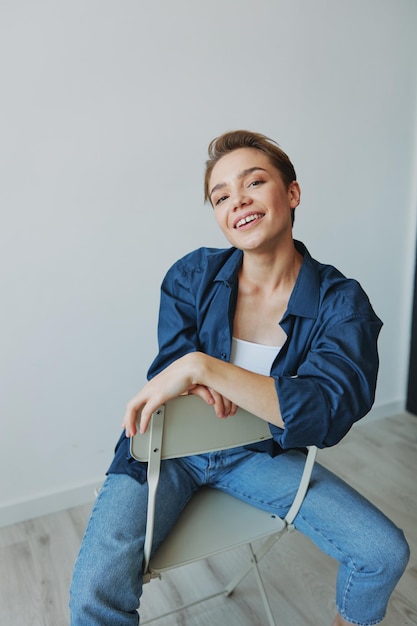 A young woman sitting in a chair at home smiling with teeth with a short haircut in jeans and a denim shirt on a white background Girl natural poses with no filters