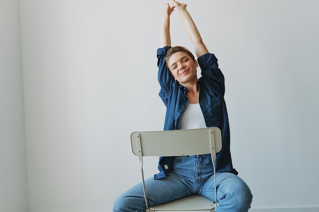 Молодая женщина сидит в кресле дома улыбается зубами с короткой стрижкой в джинсах и джинсовой рубашке на белом фоне Девушка в естественных позах без фильтров