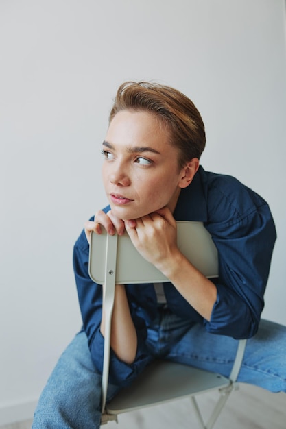 A young woman sitting in a chair at home smiling with teeth with a short haircut in jeans and a denim shirt on a white background Girl natural poses with no filters