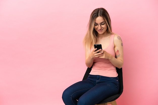 Young woman sitting on a chair over isolated pink background sending a message with the mobile