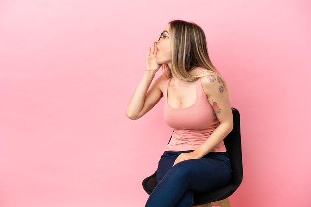 Photo young woman sitting on a chair over isolated pink background shouting with mouth wide open to the lateral