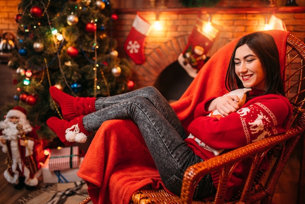 Young woman sitting on a chair near fireplace, christmas tree with decoration, xmas holiday celebration