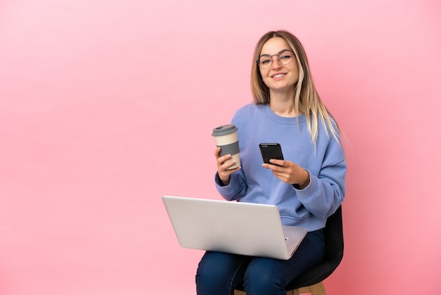 Giovane donna seduta su una sedia con un computer portatile su sfondo rosa isolato che tiene caffè da portare via e un cellulare