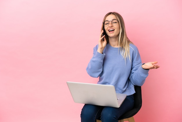 Giovane donna seduta su una sedia con un computer portatile su sfondo rosa isolato che tiene una conversazione con il telefono cellulare con qualcuno