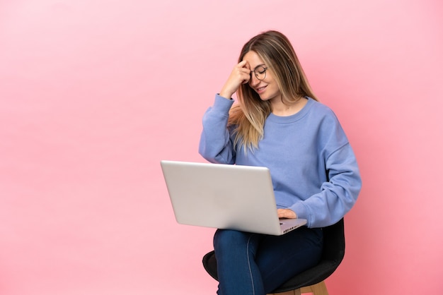 Young woman sitting on a chair with laptop over isolated pink background laughing