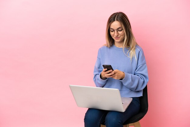 孤立したピンクの背景の上のラップトップと椅子に座っている若い女性が携帯電話でメッセージを送信します。