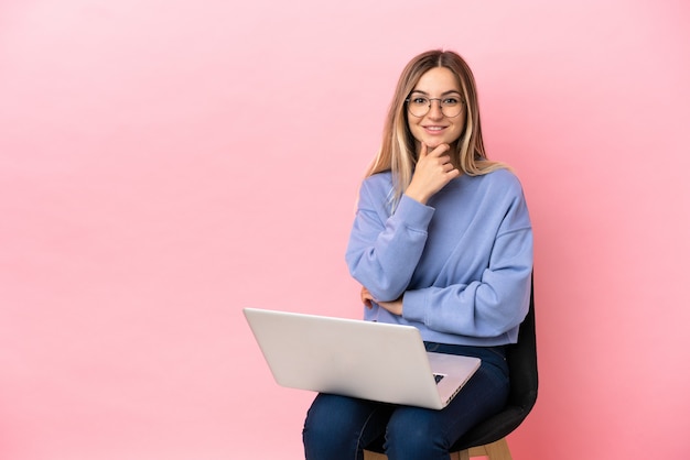 Giovane donna seduta su una sedia con laptop su sfondo rosa isolato con occhiali e sorridente