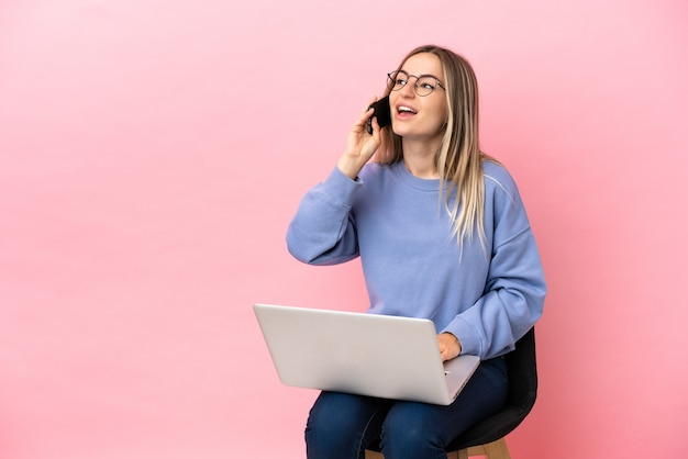 Young woman sitting on a chair with laptop over isolated pink surface keeping a conversation with the mobile phone