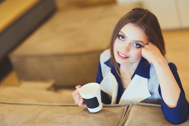 Young woman sitting on comfortable sofa with cup on coffee in hands in white room