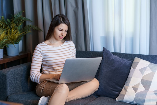 Young woman sitting on couch working at laptop browsing internet or reading news from home happy fem...