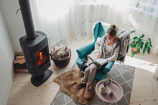 Photo young woman sitting in a cozy armchair with a warm blanket reading a book