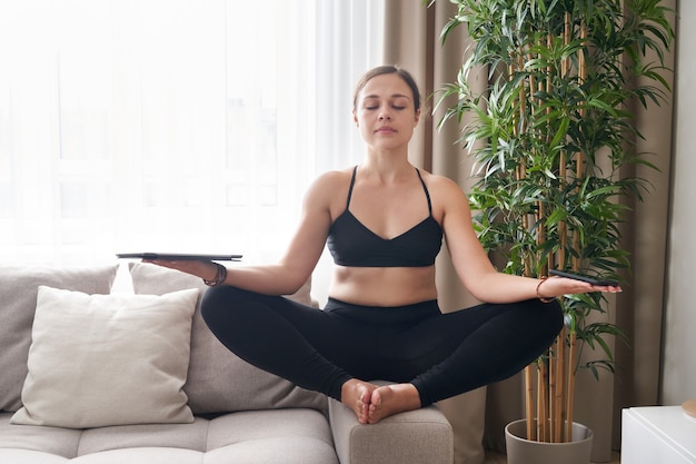 Young woman sitting crosslegged on couch in lotus pose meditating at home in living room