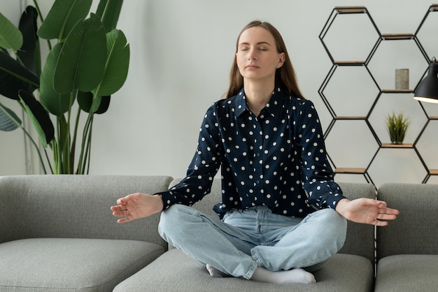 Young woman sitting crosslegged on couch with eyes closed meditating at home