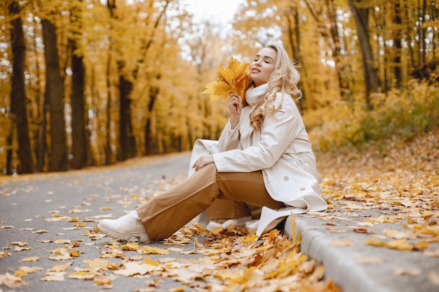 Young woman sitting on a curb in autumn forest. Blonde woman holding a yellow leaves. Girl wearing beige coat and brown trousers.