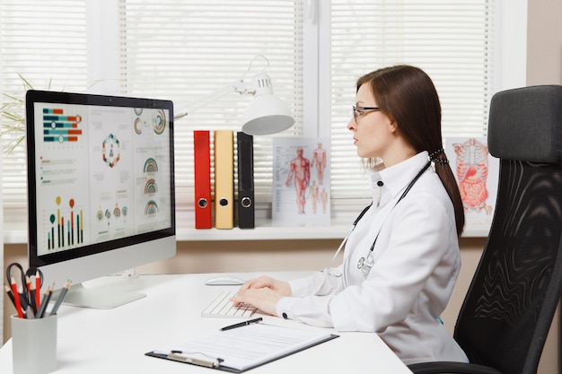 Young woman sitting at desk, working on modern computer with medical documents in light office in hospital