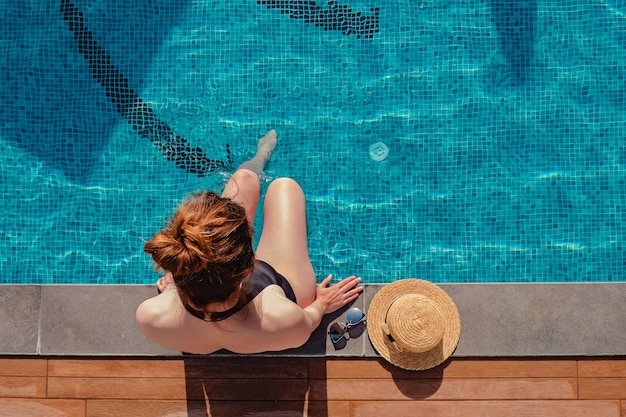 Young woman sitting on the edge of the swimming pool with her straw hat lying near top view