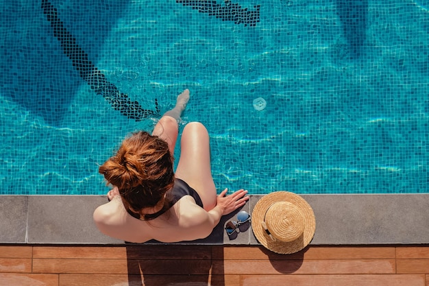 Photo young woman sitting on the edge of the swimming pool with her straw hat lying near top view