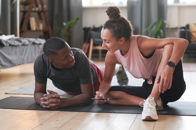 Photo young woman sitting on floor chatting to her boyfriend while he is doing plank exercise in morning