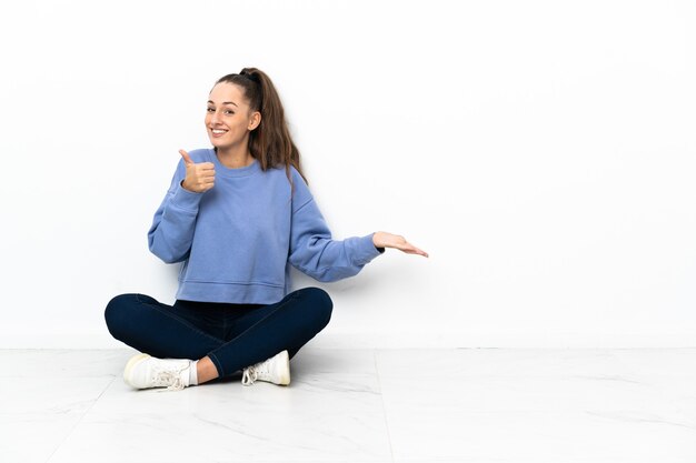 Молодая женщина сидит на полу, держа на ладони воображаемое пространство, чтобы вставить рекламу и с большими пальцами руки вверх