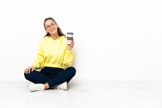 Young woman sitting on the floor holding a take away coffee looking up while smiling