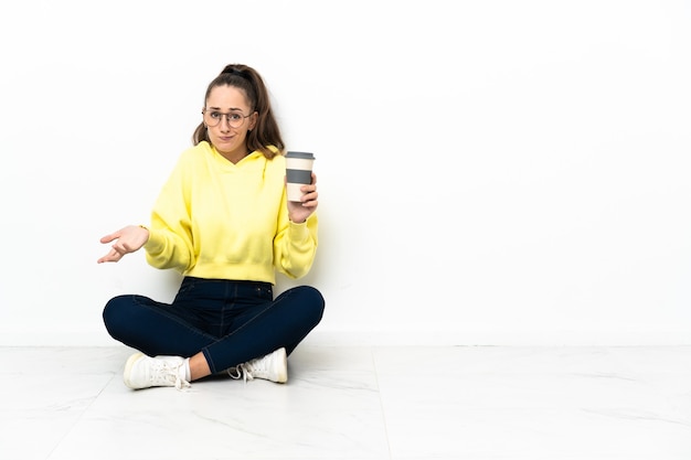 テイクアウトのコーヒーを持って床に座っている若い女性は、肩を持ち上げながらジェスチャーを疑う
