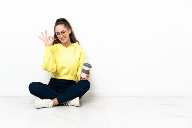 Photo young woman sitting on the floor holding a take away coffee showing ok sign with fingers