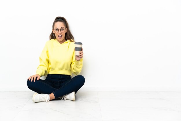 Young woman sitting on the floor holding a take away coffee with surprise and shocked facial expression