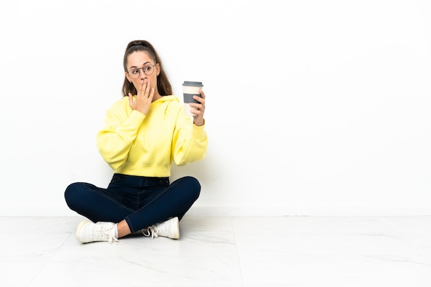 Young woman sitting on the floor holding a take away coffee with surprise and shocked facial expression