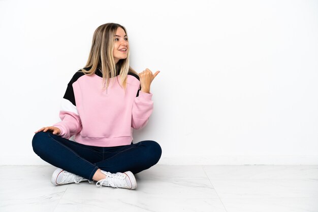 Young woman sitting on the floor at indoors pointing to the side to present a product