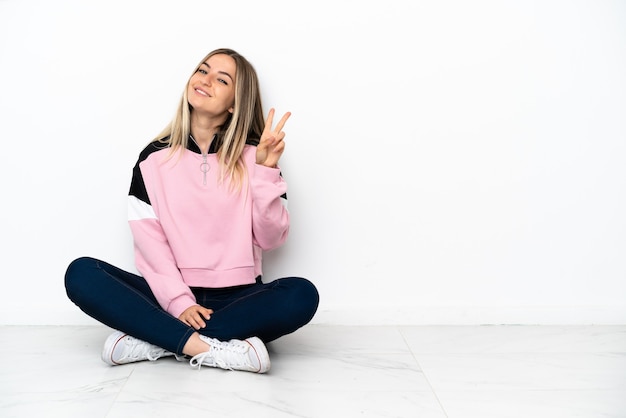 Young woman sitting on the floor at indoors smiling and showing victory sign