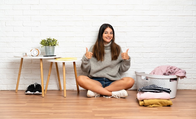 Young woman sitting on the floor at indoors with clothes basket giving a thumbs up gesture