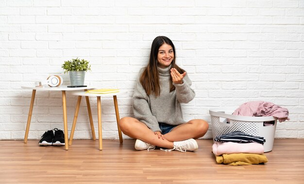 屋内の床に座っている若い女性が、手で来るように誘う洋服バスケットを持っています。あなたが来て幸せ