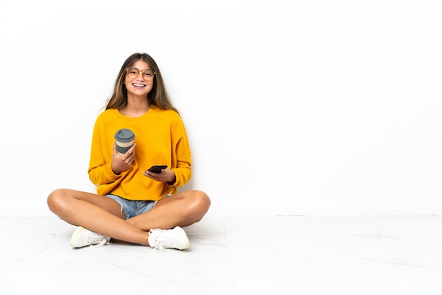 Young woman sitting on the floor isolated on white background holding coffee to take away and a mobile