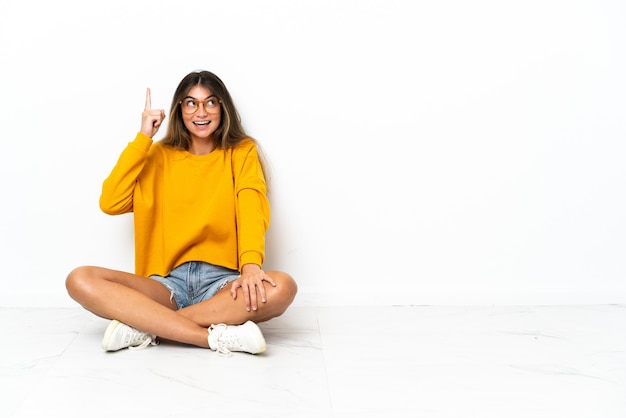 Young woman sitting on the floor isolated on white background intending to realizes the solution while lifting a finger up