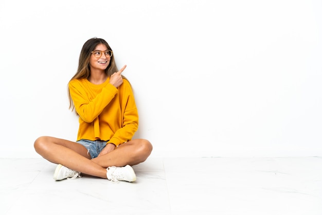 Foto giovane donna seduta sul pavimento isolata su sfondo bianco che punta il dito a lato