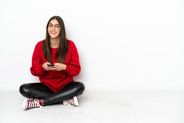 Young woman sitting on the floor isolated on white background sending a message with the mobile