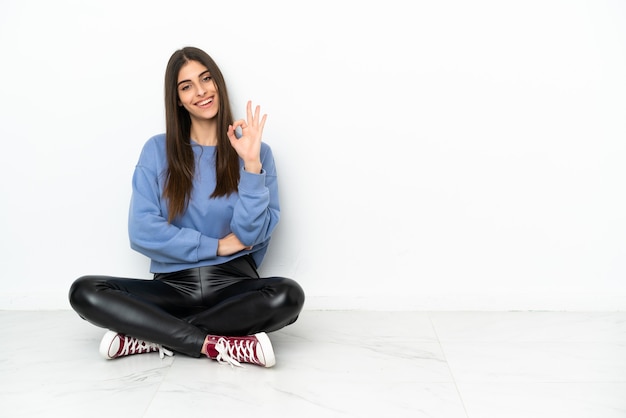 Young woman sitting on the floor isolated on white background showing ok sign with fingers