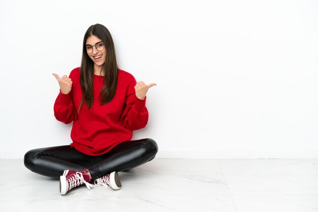 Young woman sitting on the floor isolated on white background with thumbs up gesture and smiling