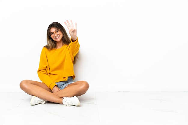 Молодая женщина сидит на полу, изолированном на белой стене, счастлива и считает четыре пальцами